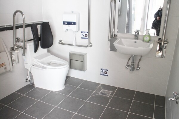 천안시 화성보건진료소 남여장애인화장실 내부는 대변기에 자동 물 내림 센서와 등받이가 양호하게 설치됐다. 비상호출벨과 휴지걸이도 대변기에 앉았을 때 손이 닿는 곳에 위치해 있다. 세면대 양쪽 손잡이는 휠체어의 세면대 접근을 용이하게 하는 상하가동식인 반면, 세면대 밑에 구조물이 있어 휠체어를 사용하는 장애인이 수도꼭지에 접근하는데 어려움이 따를 것으로 보였다. ©박종태