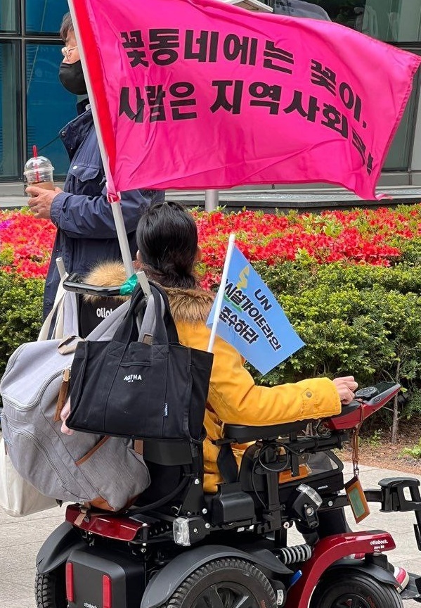 전동휠체어에 '꽃동네에는 꽃이. 사람은 지역사회로!' 현수막을 맨 중증장애인 활동가 뒷모습.ⓒ전국탈시설장애인연대
