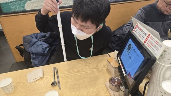 테이블오더형 무인정보단말기를 통해 주문을 하려는 시각장애인. 음성이 전혀 제공되지 않아 기기 위치나 주문 시 메뉴선택을 위한 터치를 할 수 없다.ⓒ장애인차별금지추진연대