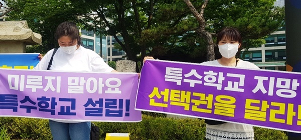 '미루지 말아요 특수학교 설립!'이 적히 보락색 현수막을 들고 서있는 여성 그 옆에 '특수학교 지망 선택권을 달라!'가 적히 보라색 현수막을 들고 있는 여성이 함께 있다